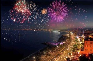Une vue nocturne d’une ville, illuminée par des feux d’artifice au-dessus d’une étendue d’eau, avec une promenade animée le long du quai et un imposant beffroi en évidence.
