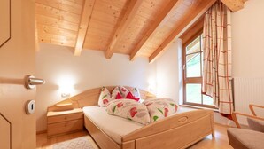A cozy bedroom with a wooden ceiling, a bed with floral bedding, a nightstand, a chair, and a window with curtains.