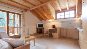 A cozy, open-plan living and kitchen area with wooden beams, a sofa, a dining table, a TV, and a kitchen counter.