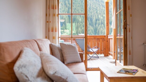 A cozy living room with a sofa, pillows, a wooden table, and a view of a forested area through large windows.
