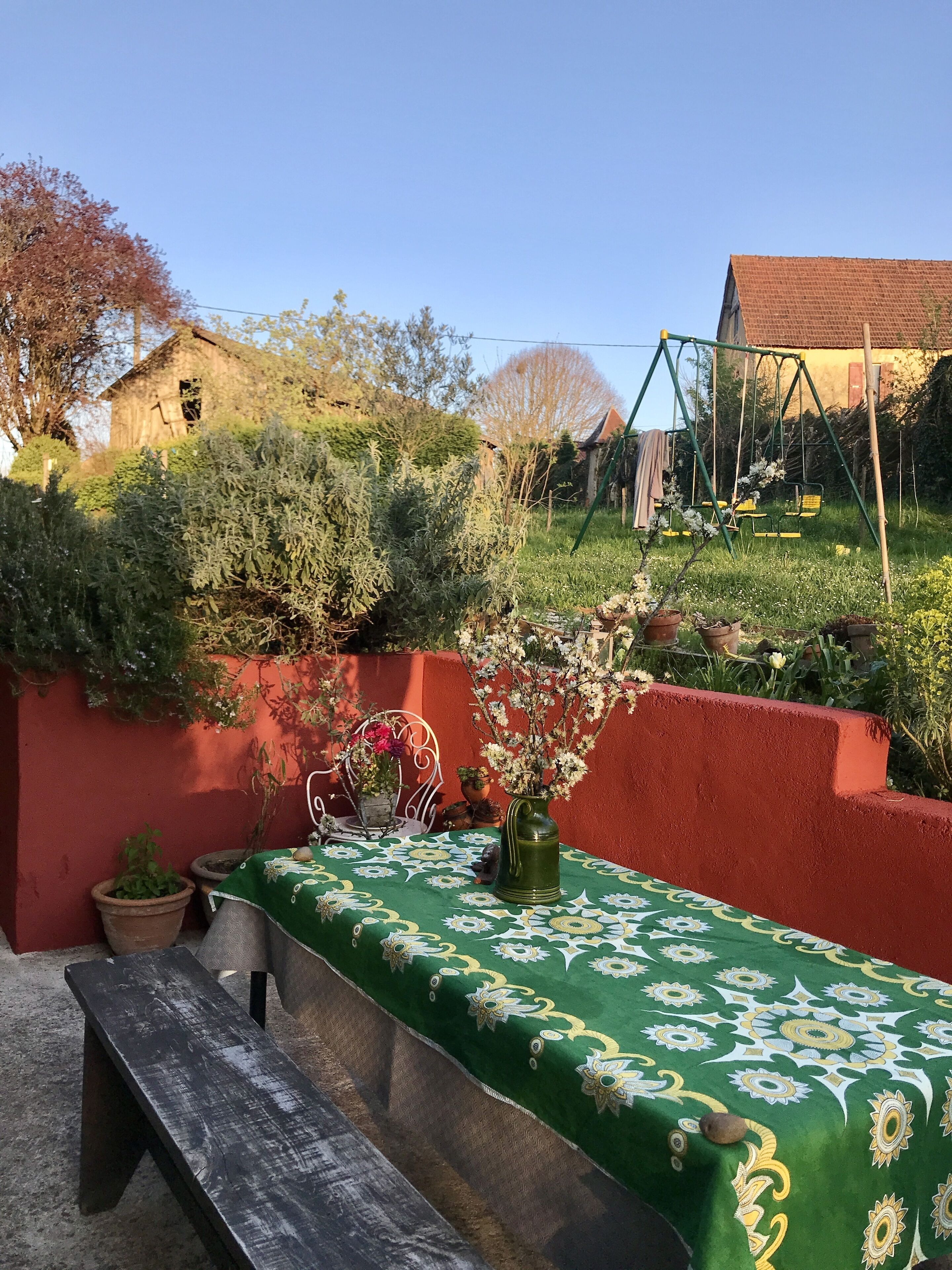 A patio with a green tablecloth, a wooden bench, potted plants, and a swing set in the background.
