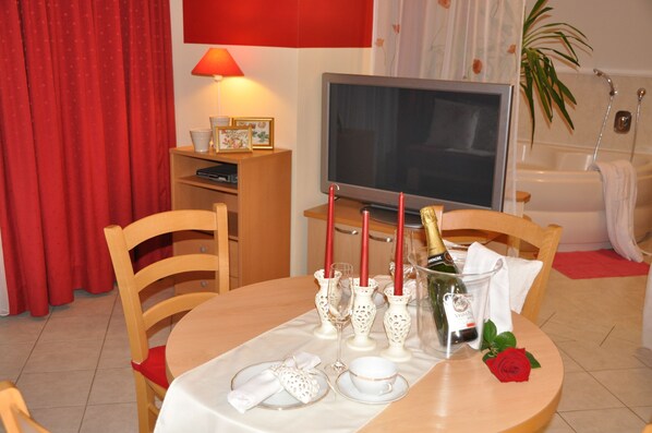 A dining area with a table set for two, a television, and a bathtub in the background.