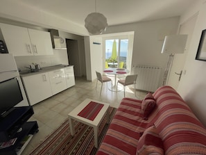 A compact living space with a kitchenette, a dining area with a table and chairs, a sofa, and a view through a large window.
