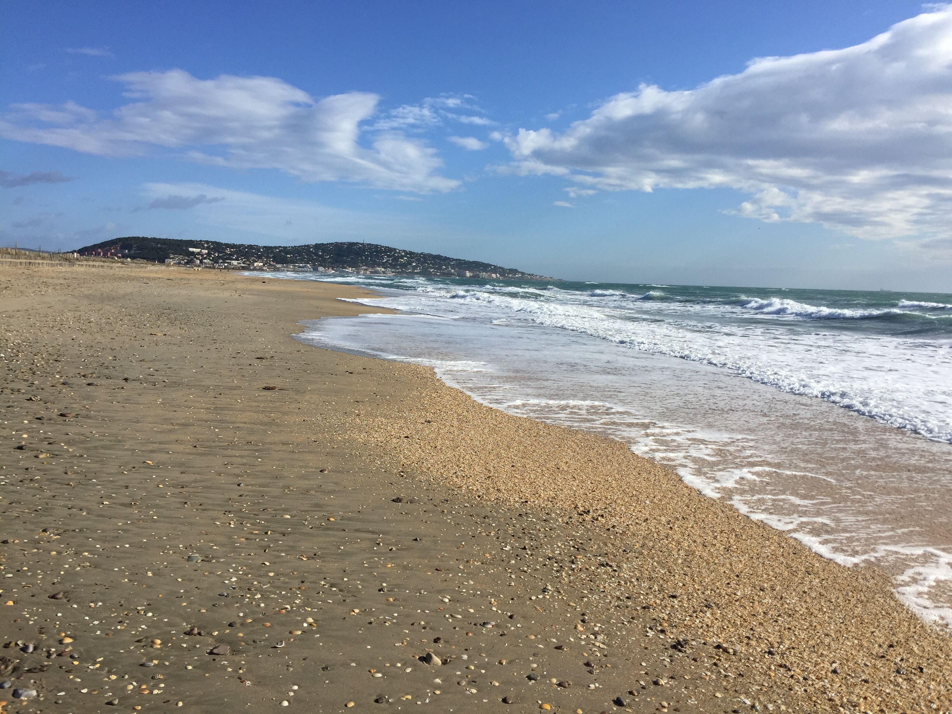 Une plage de galets, des vagues, un ciel dégagé et une colline qui se dessine à l’horizon.