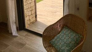 Une pièce avec un mur en pierre, un fauteuil en osier doté d’un coussin, et une vue sur un paysage verdoyant, avec des arbres et des collines.