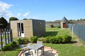 A small wooden building with a door and window, a stone wall, and a fenced area with a lawn and shrubs.