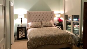 A bedroom with a tufted headboard, a bed with patterned bedding, two bedside tables with lamps, a ceiling fan, and a window with a view of the outside.