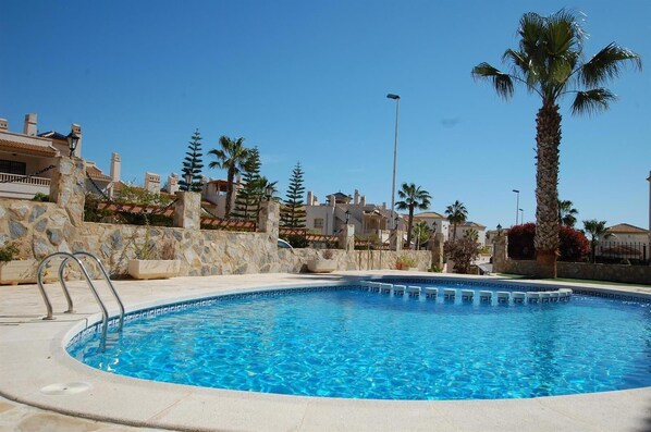 A clear blue swimming pool with a stone wall and palm trees, surrounded by buildings with Mediterranean architecture. - Beautiful apartment 2ch 2sdb Las Ramblas Golf Orihuela (villacosta)