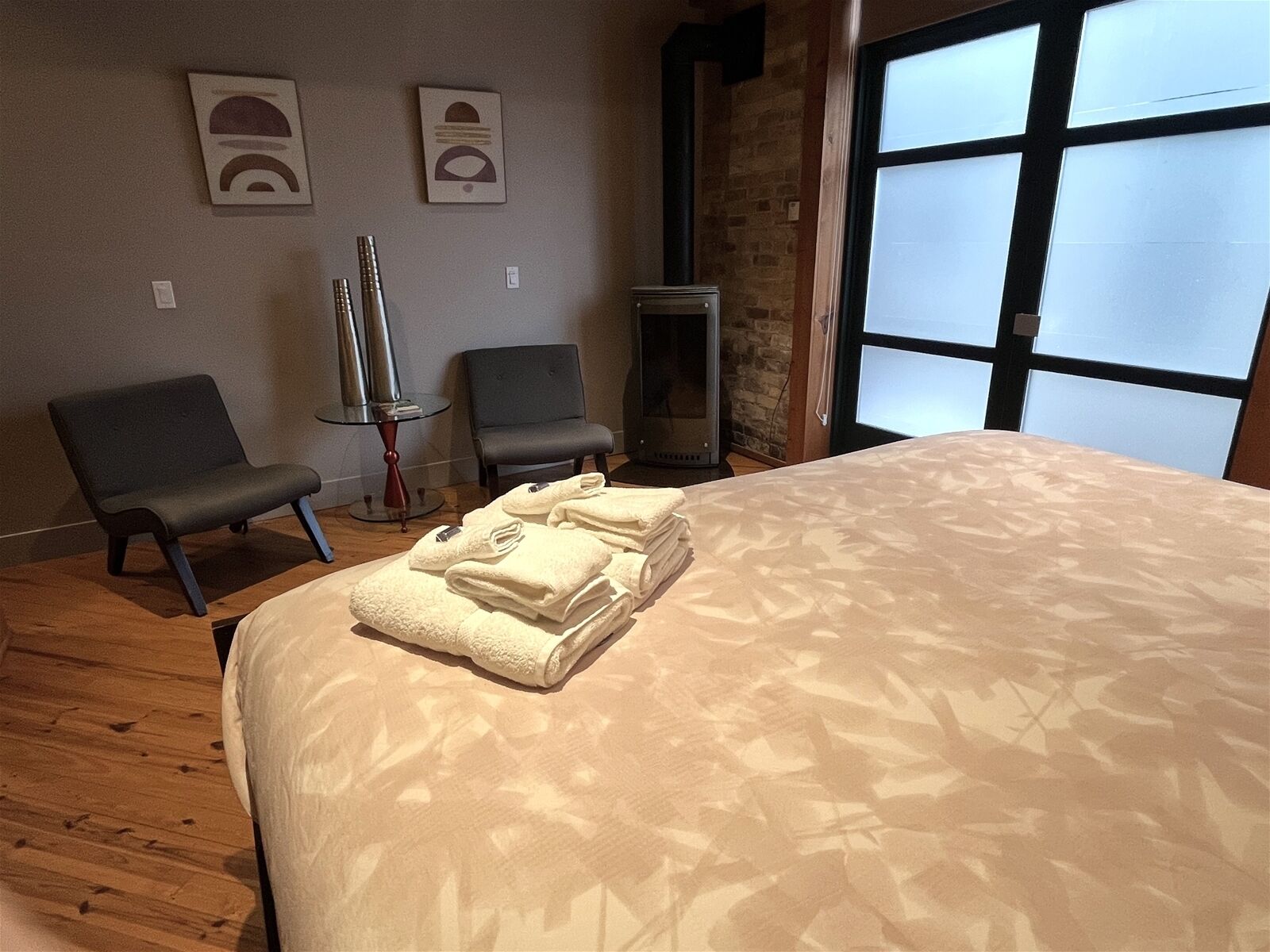 A hotel room with a large bed, two armchairs, a small table with a lamp, and a fireplace.
