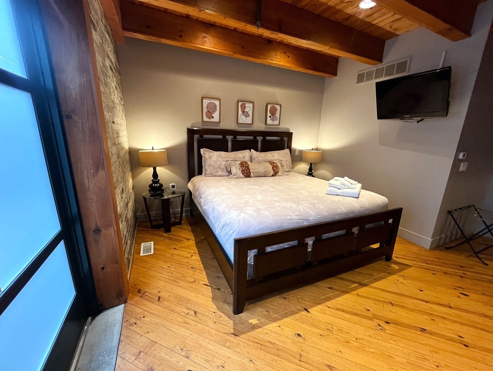A bedroom with a wooden bed, two bedside tables with lamps, a flat-screen TV mounted on the wall, and a window with a view of the sky.