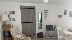 Ein Wohnzimmer mit einer Couch, einem Sessel und einem Esstisch mit Stühlen. Ein Fernseher steht auf einem Beistelltisch, eine Lampe ist vorhanden und es gibt ein Bücherregal.