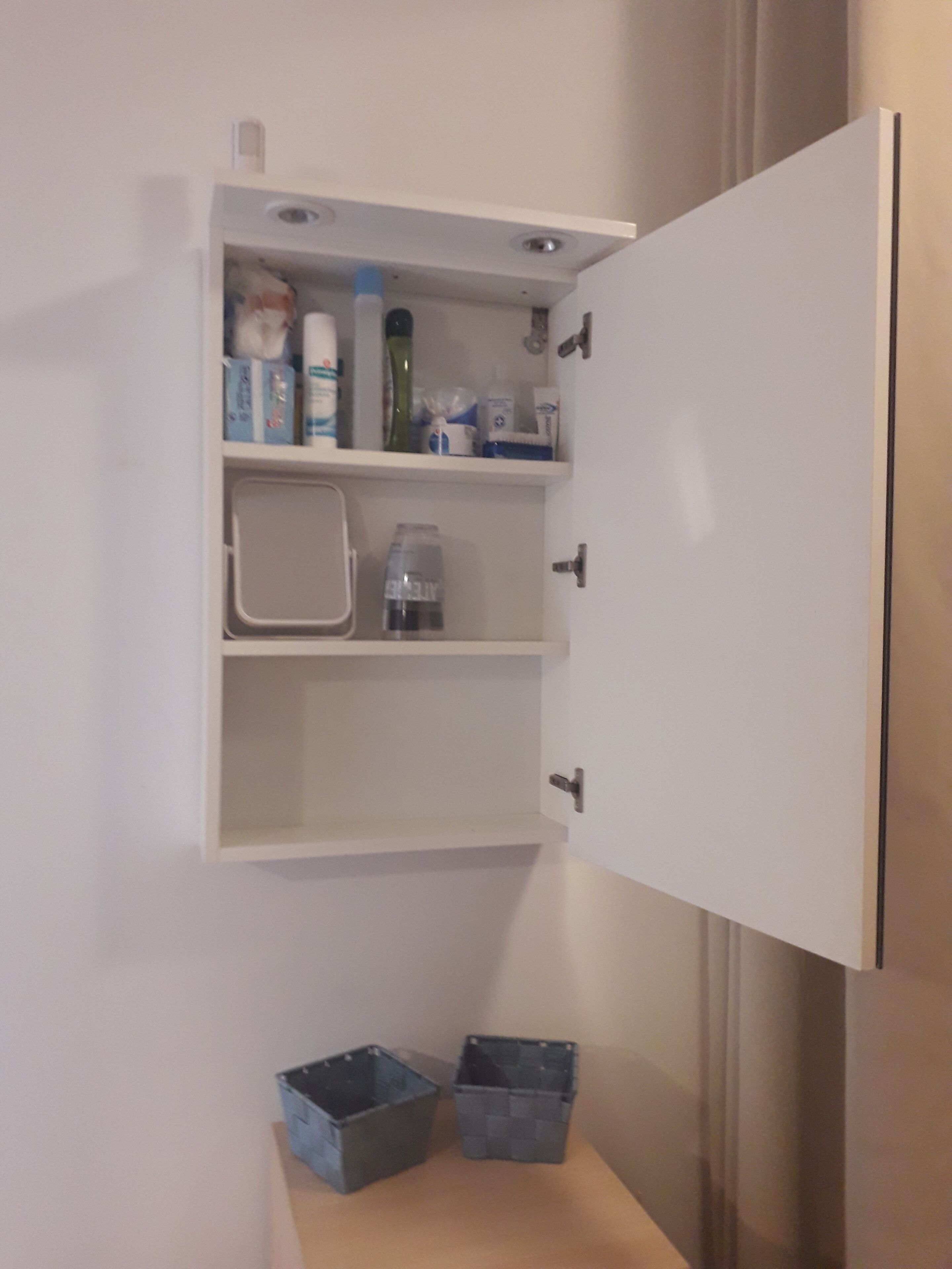 A white cabinet with a shelf containing toiletries and a bottle of water. There are two blue bins on the floor.