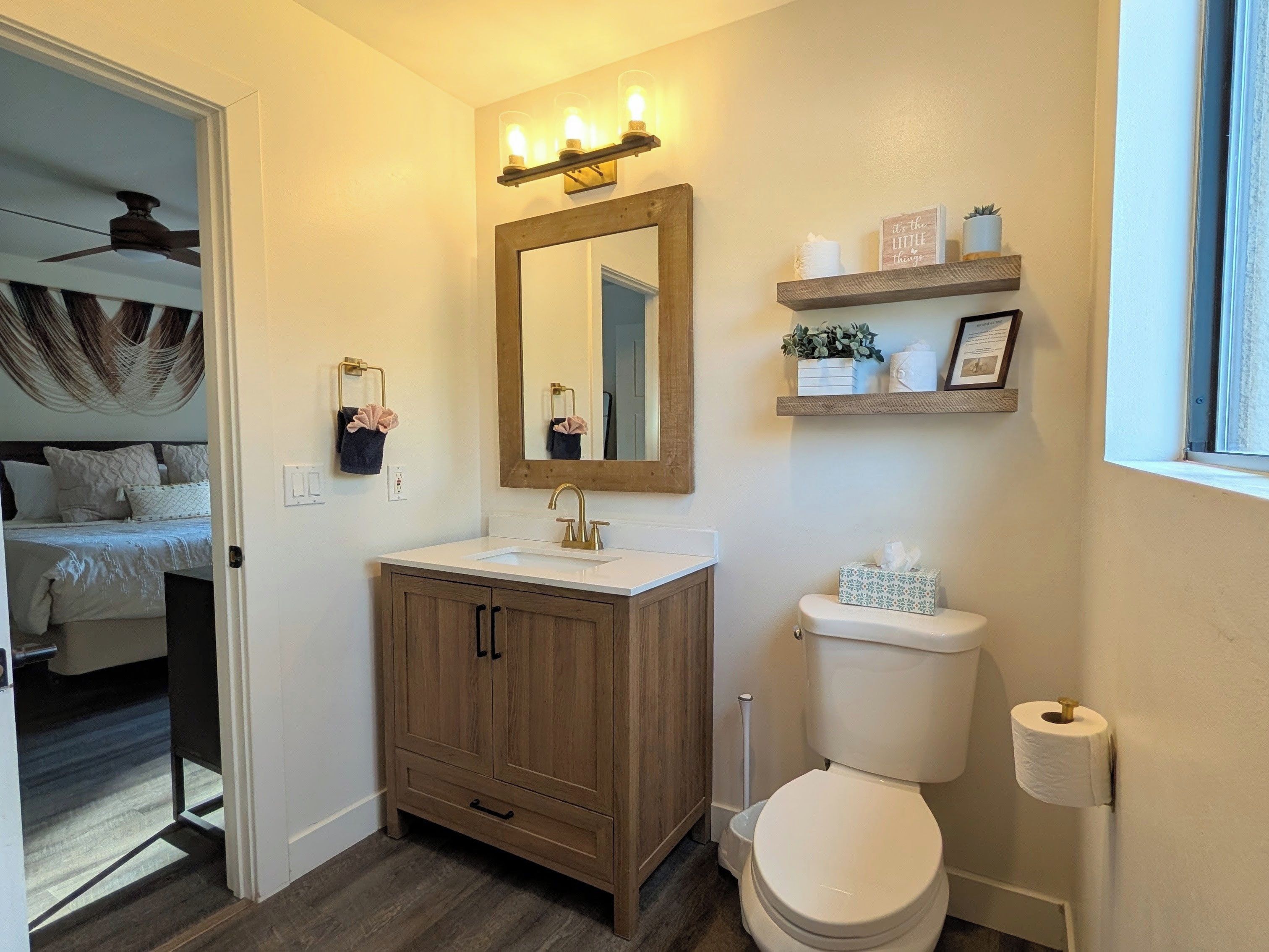 A bathroom with a wooden vanity, a white sink, a mirror, and a toilet. There is a window with a view of the outside.