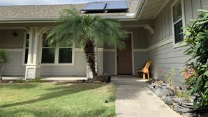 Una casa con un porche cubierto, paneles solares en el techo, una palmera y un pequeño jardín con rocas y plantas.