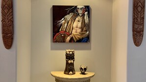 Un pasillo con una mesa pedestal central, una obra de arte enmarcada encima y dos esculturas de madera en las paredes.