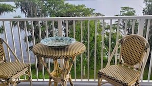 Deux chaises en osier et une petite table ronde sont disposées sur un balcon doté d’une balustrade blanche, d’où l’on peut admirer un plan d’eau et des arbres.