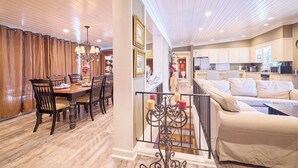 Un coin repas avec une table et des chaises en bois, un lustre et un escalier menant à un espace de vie comprenant un canapé et une cuisine en arrière-plan.