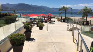 A terrace with potted plants, red umbrellas, and a view of the beach and mountains. - OCEANFRONT CONDO IN TROPICAL PARADISE AWAITS YOU AT THE GRAND VENETIAN! (Las Glorias)