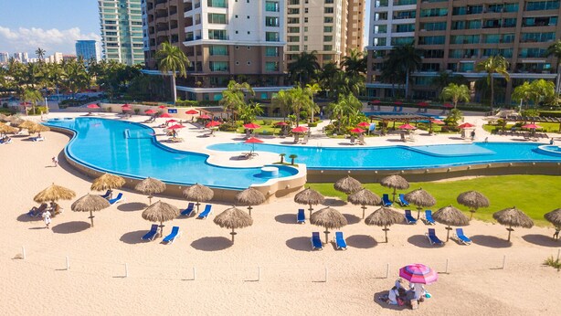 A resort with a large swimming pool, sun loungers, and thatched umbrellas. - OCEANFRONT CONDO IN TROPICAL PARADISE AWAITS YOU AT THE GRAND VENETIAN! (Las Glorias)