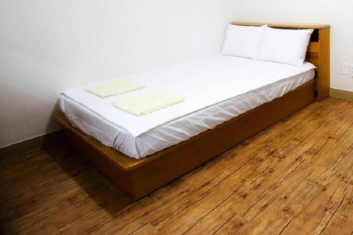 Un lit simple avec une tête de lit en bois, dans une pièce dont le sol est recouvert de planches de bois et les murs sont peints en blanc.