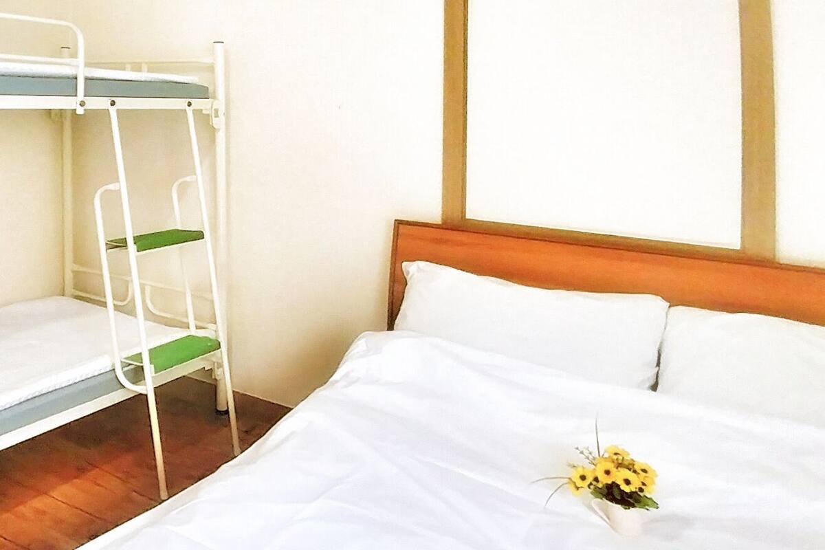 Un lit simple avec une literie blanche et un petit arrangement floral jaune. Un lit superposé est placé sur le côté gauche.