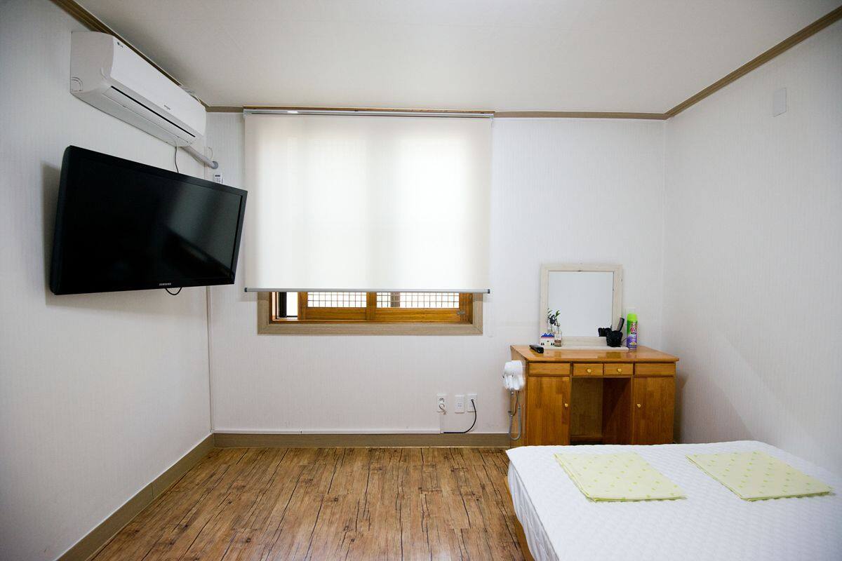Une chambre avec un téléviseur fixé au mur, une commode en bois, une fenêtre avec des stores et un lit recouvert d’une couverture verte.