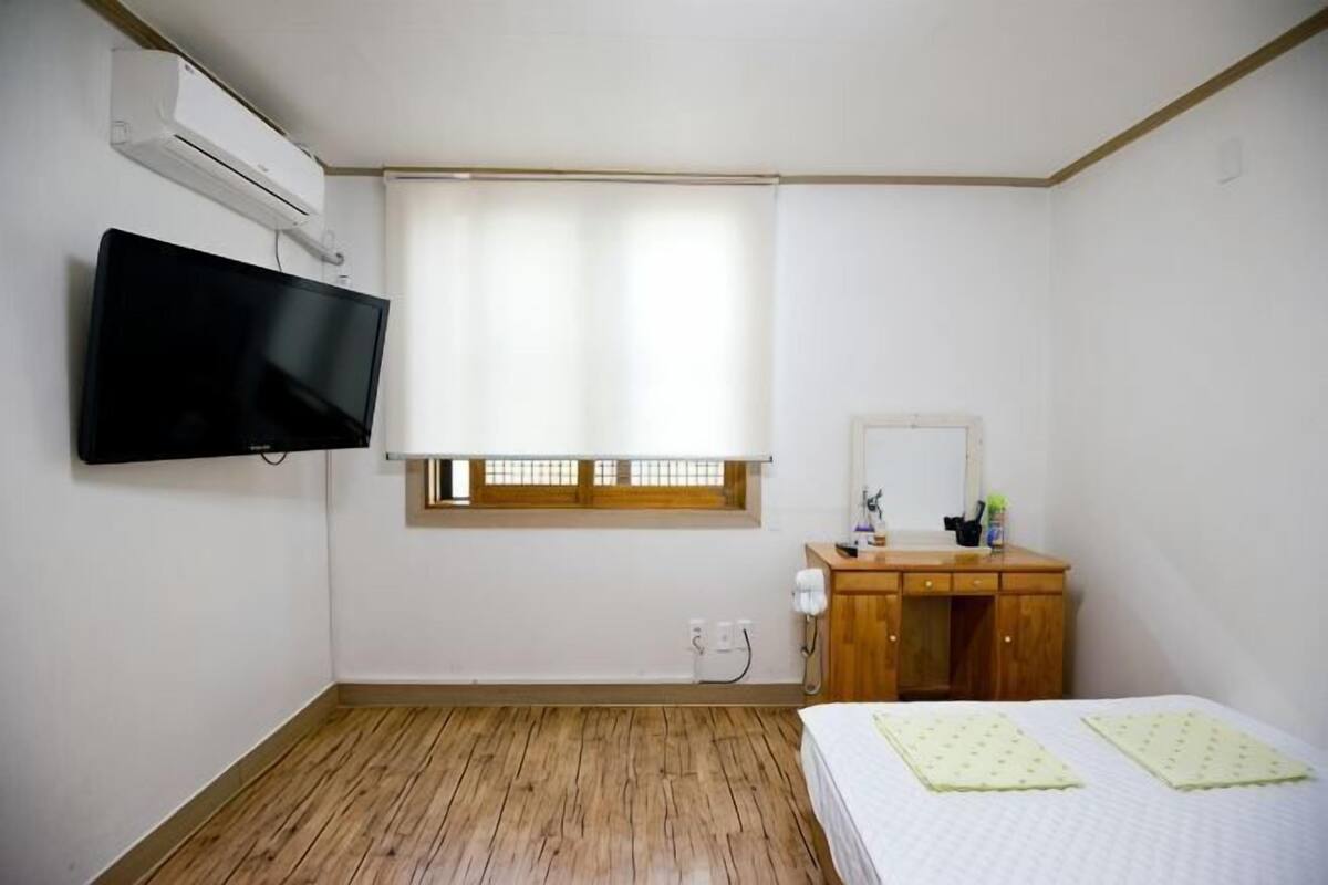 Une chambre à coucher comprenant un lit, une télévision, une fenêtre avec des stores, une commode en bois et un miroir.