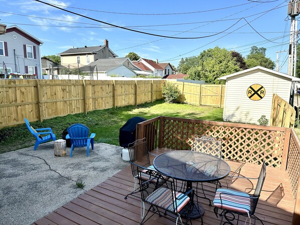 A backyard with a wooden deck, blue chairs, a round metal table, and a shed with a railroad crossing sign. - Walk to Downtown - 10 Mins to Starved Rock! (Lasalle)