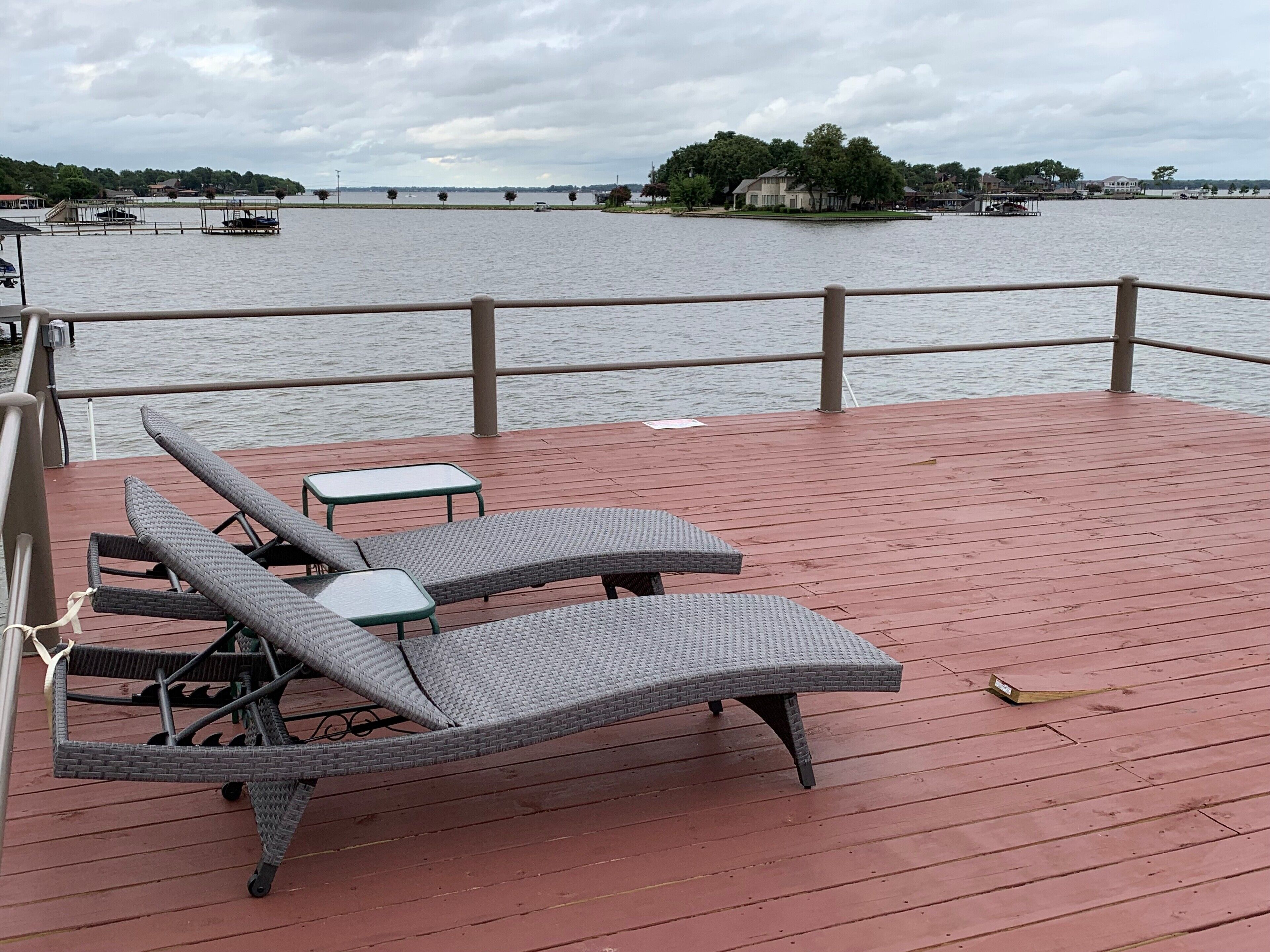 Une terrasse dotée de fauteuils de jardin en osier et d’une petite table, offrant une vue sur une étendue d’eau calme, avec des bateaux et des maisons visibles au loin.