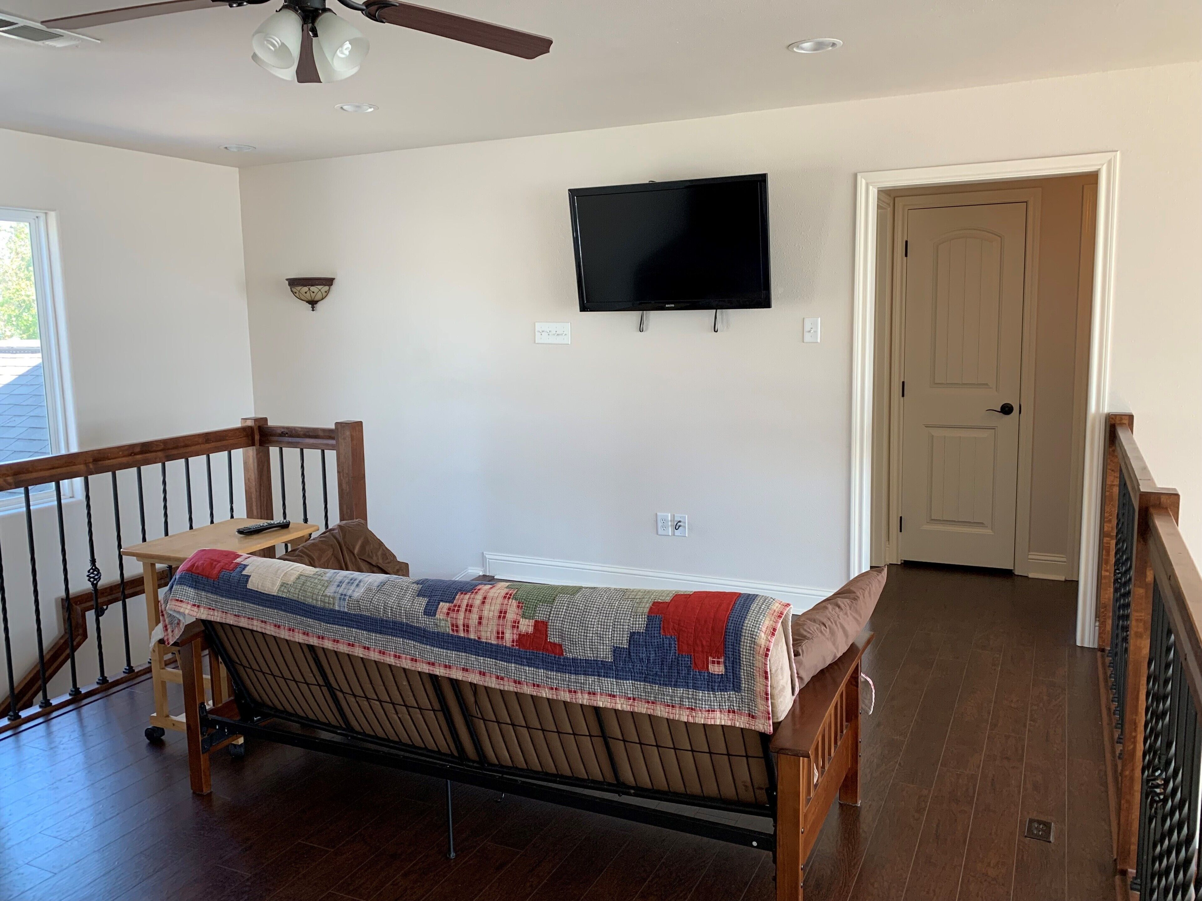 Una habitación con una cama de madera, un ventilador de techo, un televisor montado y una puerta.