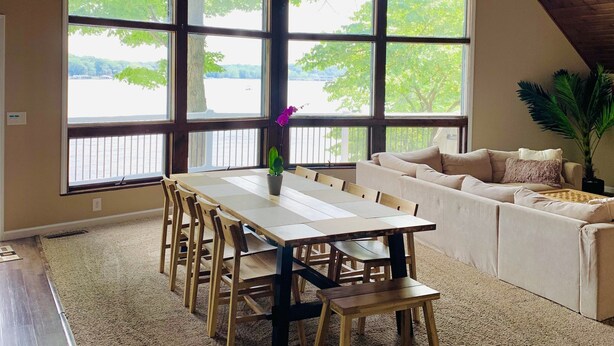 Ein großzügiger Raum mit hoher Decke, großen Fenstern mit Blick auf Bäume und ein Gewässer, einem Esstisch mit Stühlen und einem Sofa.
