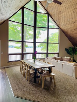 Una habitación amplia con techo alto, ventanas grandes que ofrecen vistas a árboles y una masa de agua, una mesa de comedor con sillas y un sofá.