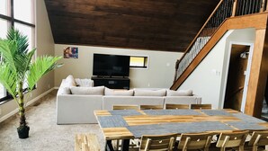 Ein Wohnzimmer mit einer Eckcouch, einem Fernseher, einem Esstisch mit Stühlen, einer Treppe und einem großen Fenster.