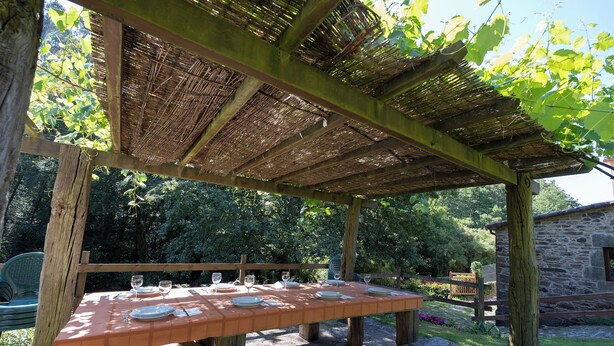 An outdoor wooden pergola with a table set for dining, surrounded by greenery and a stone building.