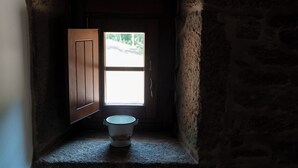 A narrow, stone-walled hallway with a wooden door and a window. A white bucket is placed on a ledge at the end of the hallway.