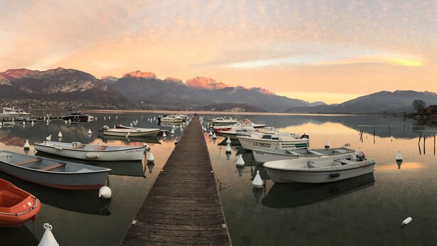 A calm lake with boats docked at a pier, surrounded by mountains and a clear sky.