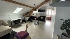 A loft-style room with a sloped ceiling, a desk with a chair, a kitchenette, a sofa, and a large plant.