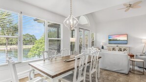 A dining area with a wooden table and white chairs, large windows with blinds, a ceiling fan, and a TV on the wall. - Pet Friendly ~ Community Pool (Miramar Beach)