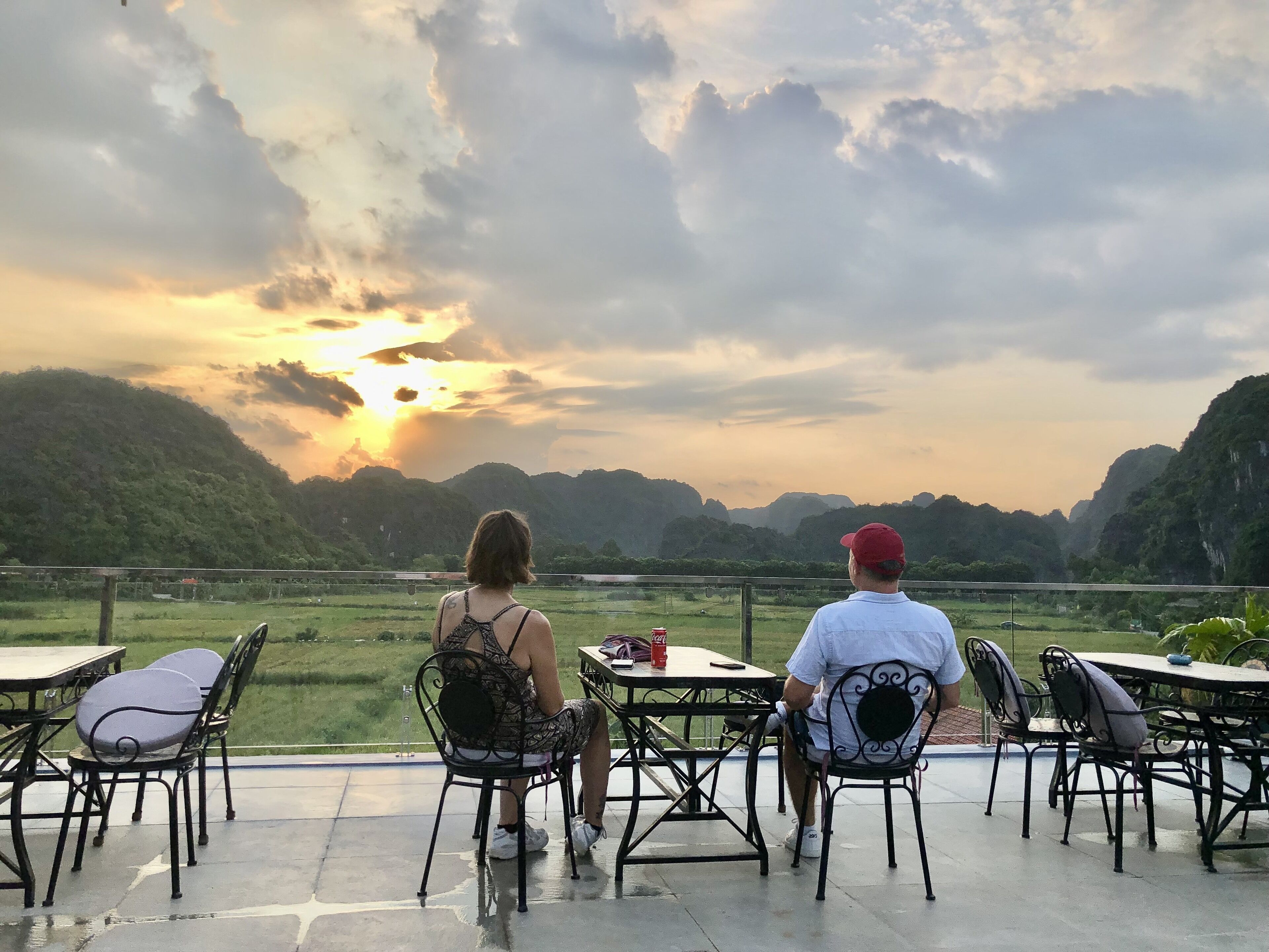 Dos personas sentadas en una mesa con una vista panorámica del atardecer sobre un paisaje montañoso.