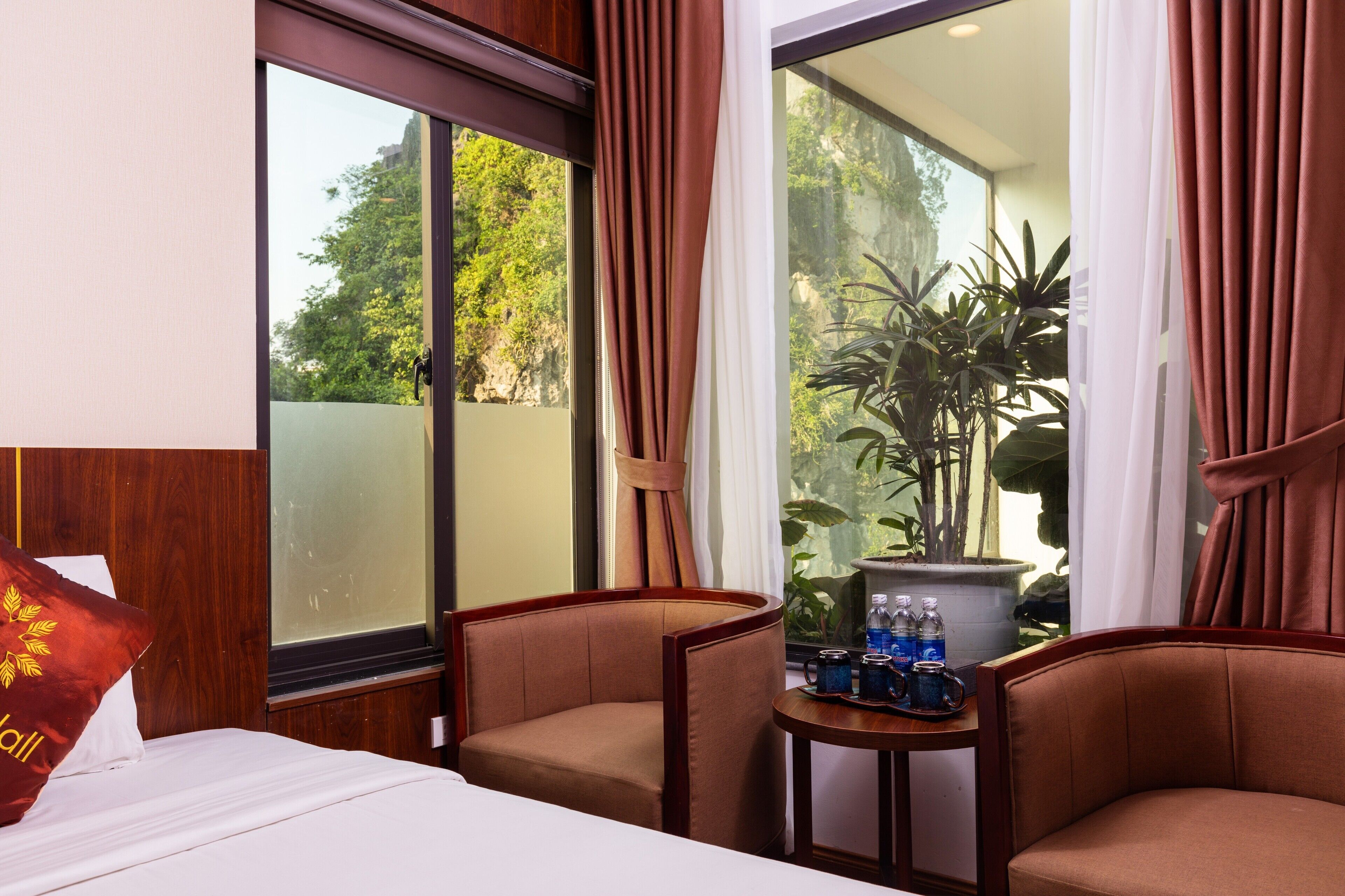 A hotel room with a bed, two armchairs, a small table with water bottles, and a view of greenery outside.