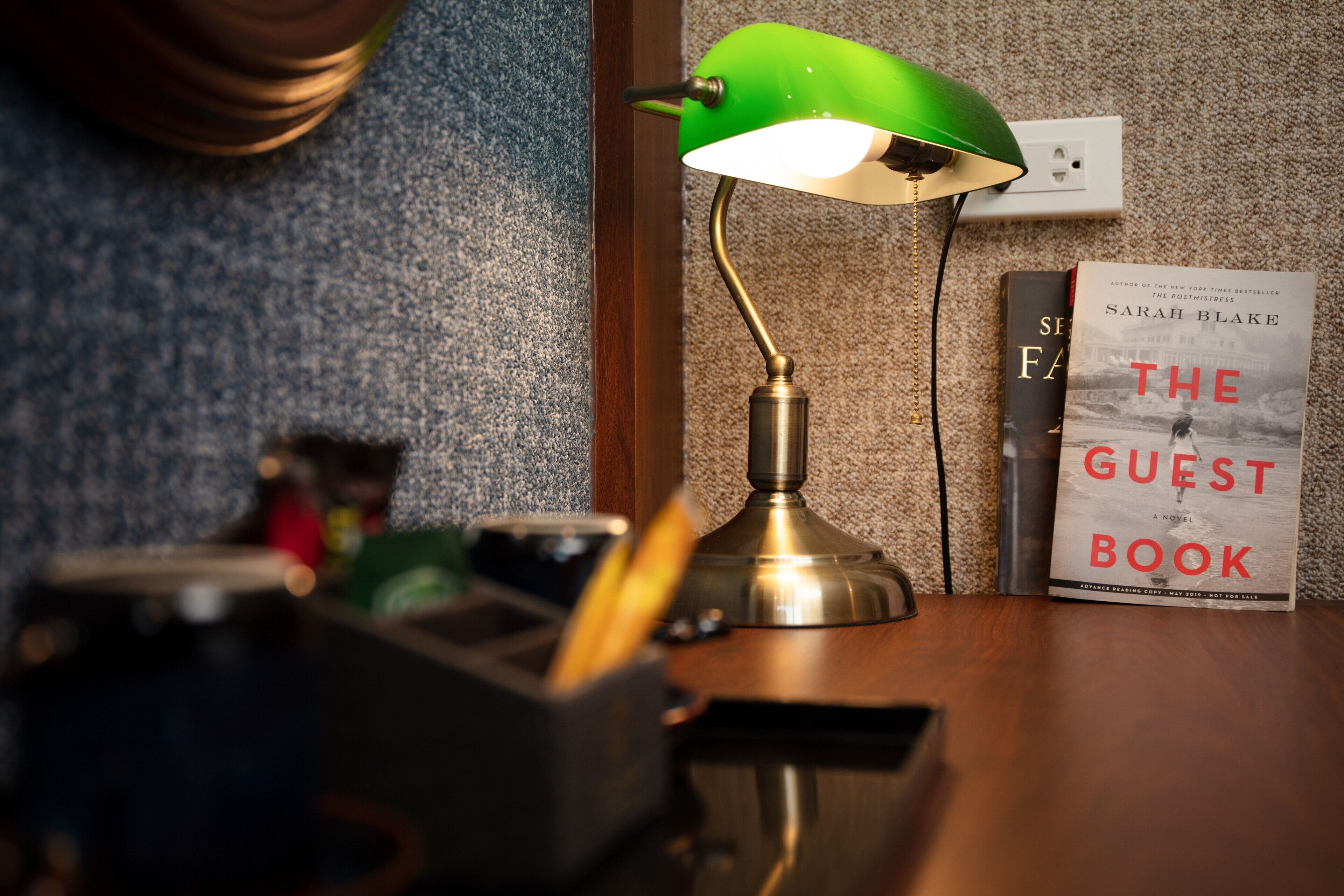 A desk with a green desk lamp, a book titled "The Guest Book" by Sarah Blake, and a pen holder.