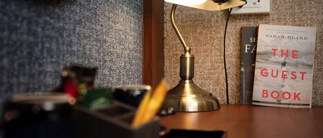 A desk with a green desk lamp, a book titled "The Guest Book" by Sarah Blake, and a pen holder.