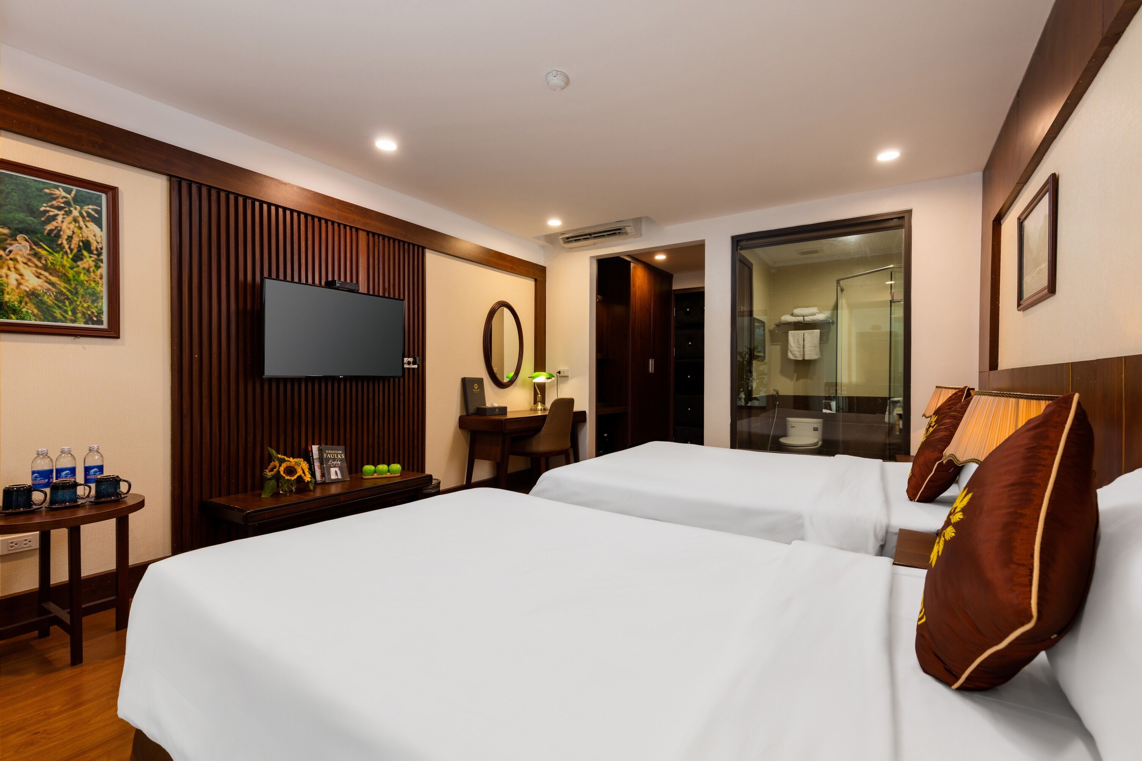 A hotel room with two beds, a TV, a desk with a chair, a small table with water bottles, and a bathroom visible through an open door.