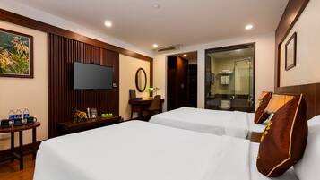 A hotel room with two beds, a TV, a desk with a chair, a small table with water bottles, and a bathroom visible through an open door.