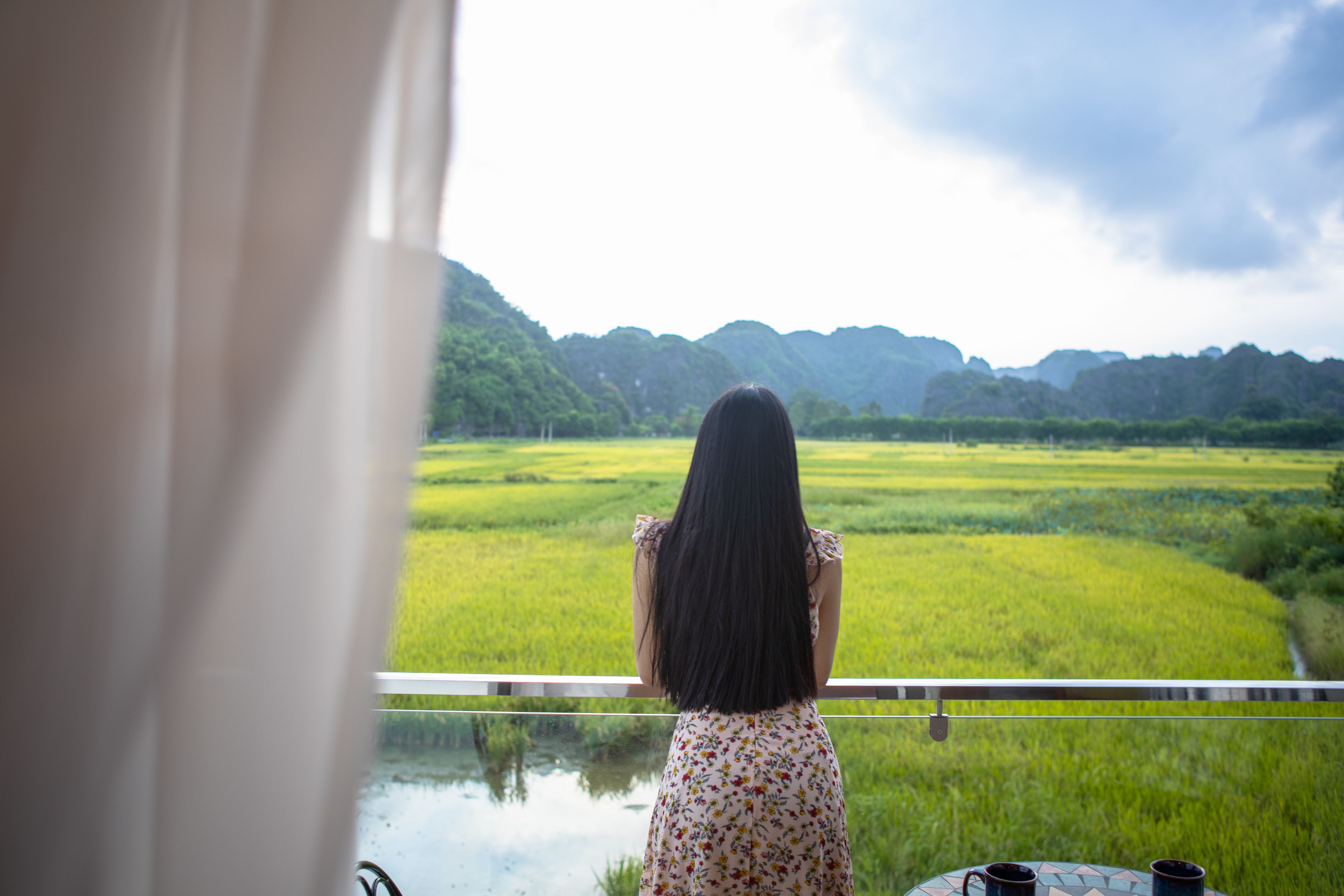 Una mujer con un vestido floral de pie junto a una ventana que da a un paisaje verde con colinas.