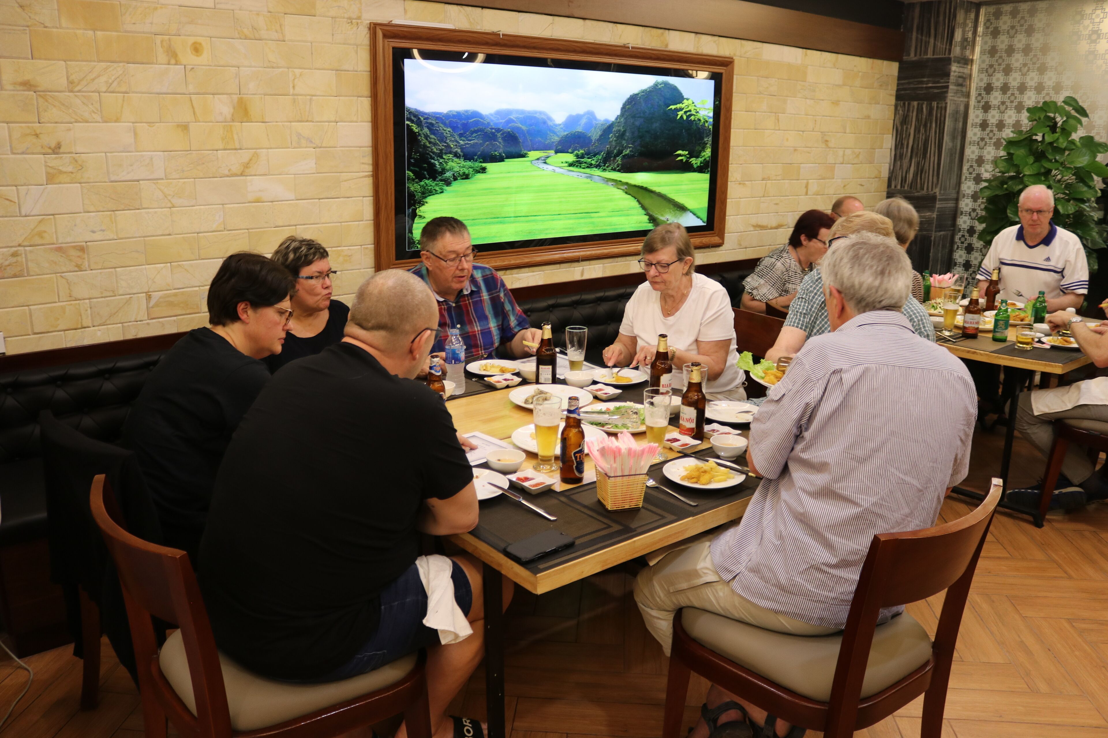 A group of people seated around a table in a room with a large TV screen displaying a landscape.