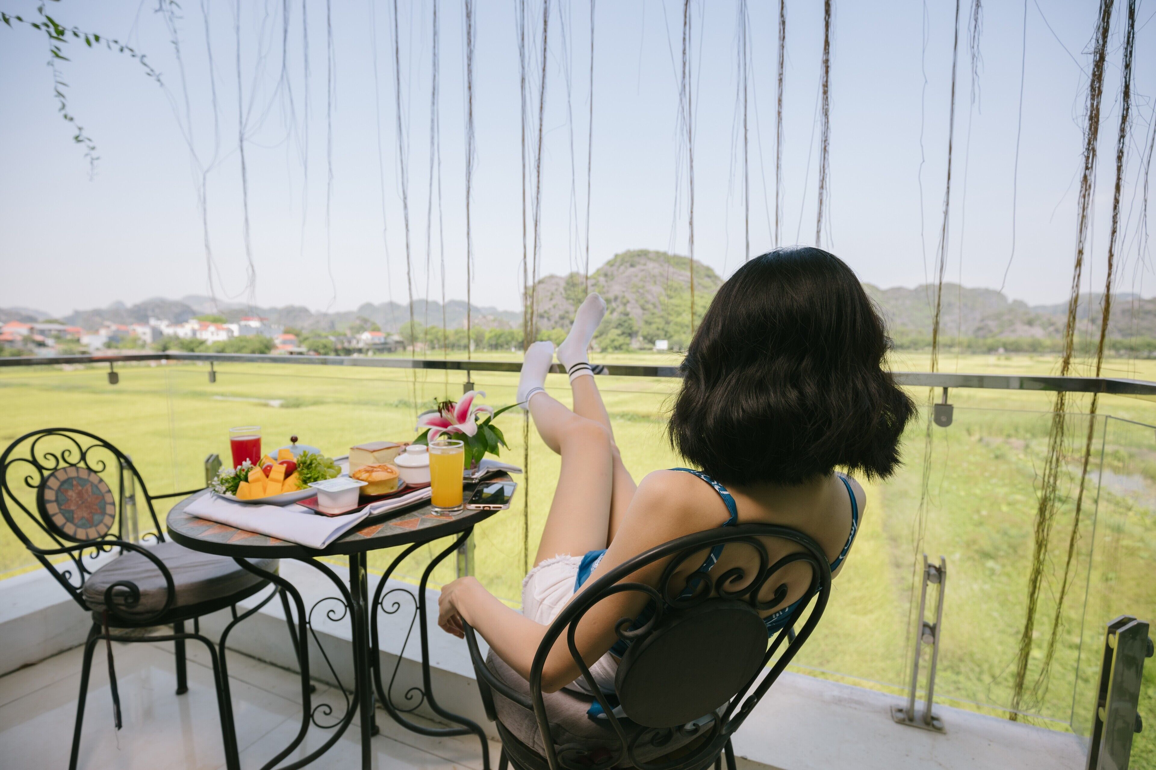 A woman sitting at a table with a view of a grassy field and hills in the background.