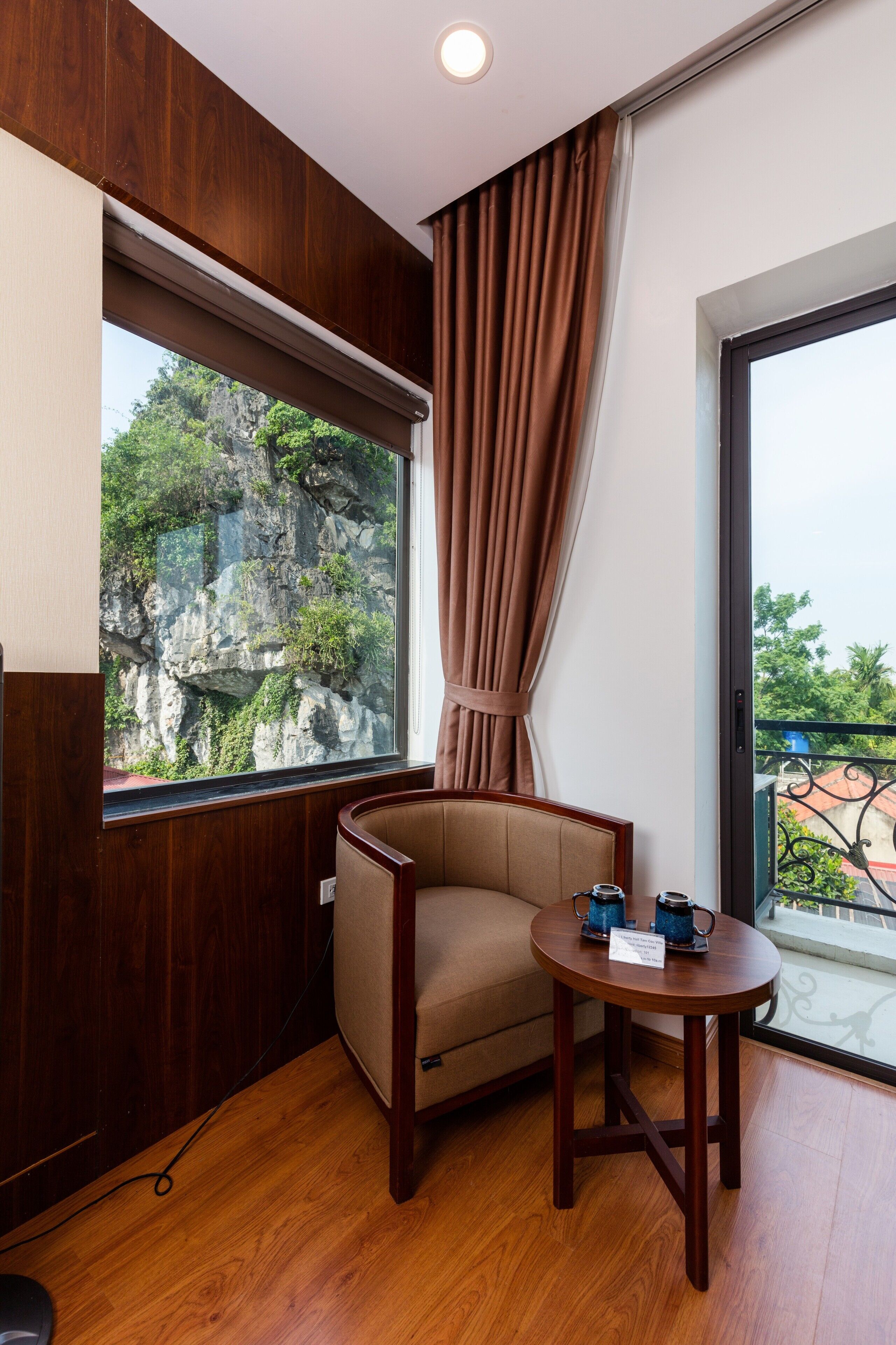 A corner room with a wooden wall, a window with a view of rocky cliffs, a beige armchair, a small round table with two cups, and a balcony with a railing.