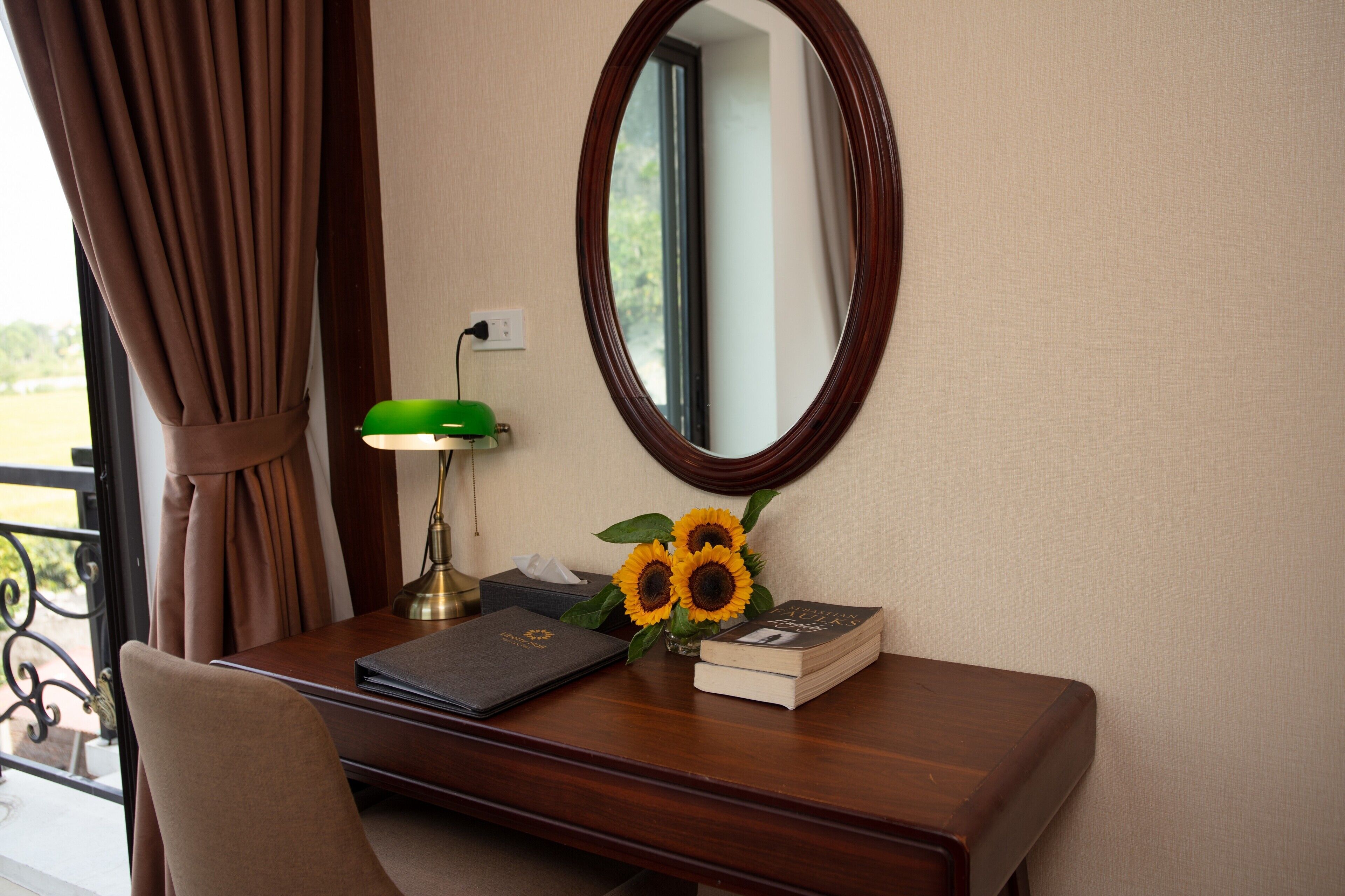 A wooden desk with a mirror, a lamp, a book, and a vase with sunflowers.