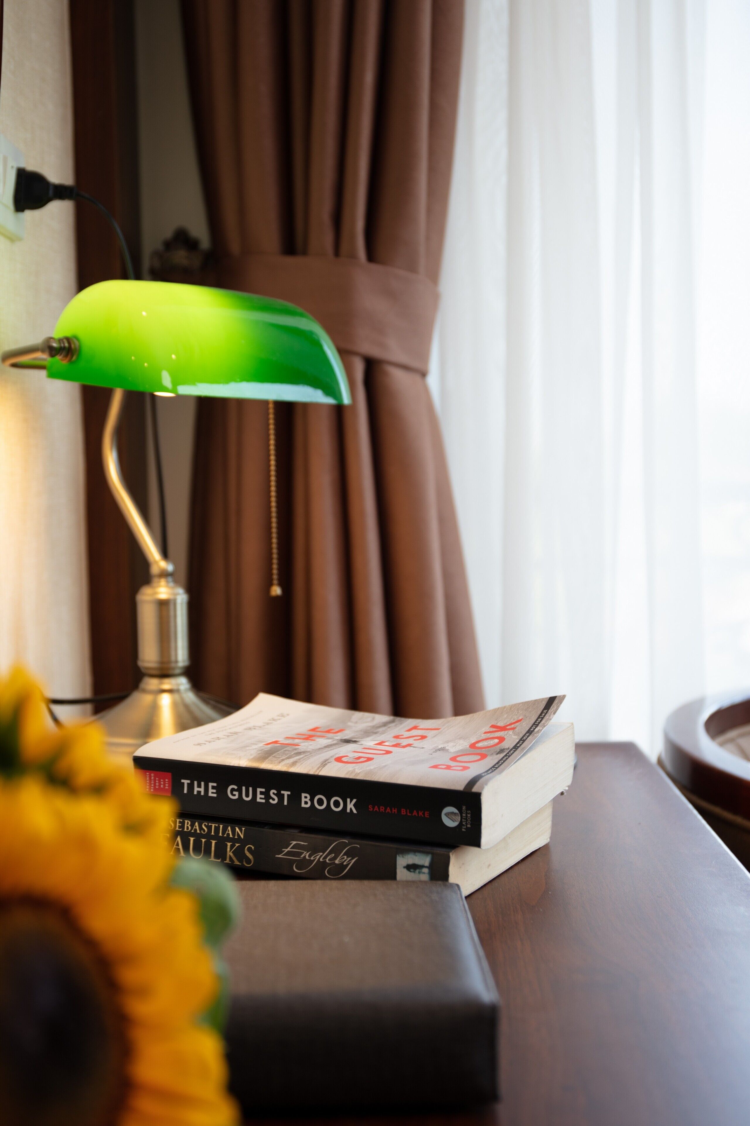 A stack of books on a wooden surface with a green desk lamp and a sunflower in the foreground.